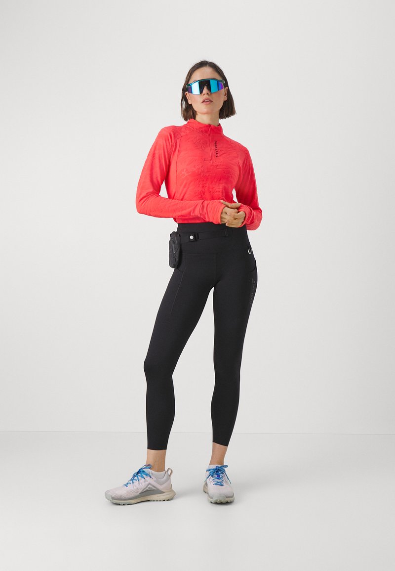 Haut actif rouge à manches longues avec demi-fermeture éclair, leggings noirs taille haute, et chaussures de sport blanches avec des accents bleus, plus une pochette noire.