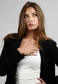 Modelo femenina con un blazer negro sobre una blusa blanca, con cabello largo castaño, usando un anillo de oro, posando con una mano en el pecho.