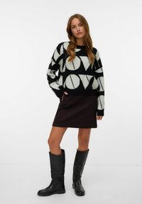 Maglione a maniche lunghe con motivo in bianco e nero e forme geometriche audaci, abbinato a una gonna marrone scuro e stivali neri in pelle al ginocchio.