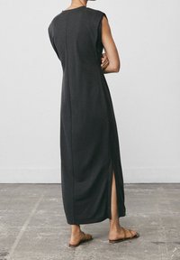 Kvinna står med armarna i kors iförd en ärmlös mörkgrå maxiklänning med sidoklipp och bruna platta sandaler på betonggolv.