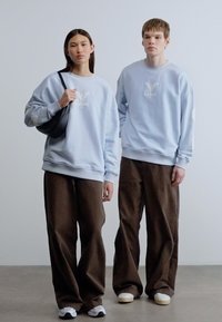Ανοιχτό γαλάζιο φούτερ συνδυασμένο με καφέ παντελόνια φαρδιάς γραμμής. Τα φούτερ έχουν μικρό κεντημένο λογότυπο. Υφή υφάσματος με άνετη εφαρμογή.