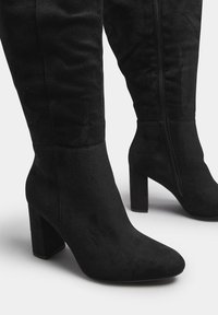 Czarne zamszowe kozaki do kolan z grubym obcasem, spiczastym noskiem i bocznym zamkiem. Gładka tekstura i dopasowany krój.