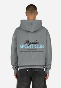 Šedá bavlnená mikina s kapucňou na stiahnutie. Na chrbte je modrý a čierny text: "Pegador SPORT CLUB WHISPERS OF COLD." Minimalistický dizajn.