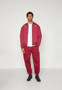 Vörös sportos dzseki és nadrág szett sima anyagból, rugalmas mandzsettákkal, cipzáras záródással és logó részletekkel. Fehér inggel viselve.