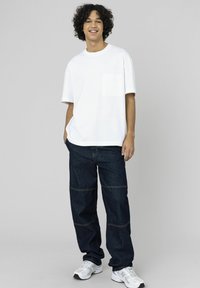 T-shirt blanc à manches courtes avec une poche poitrine, associé à un pantalon large en denim foncé avec des surpiqûres contrastantes et des coutures visibles.