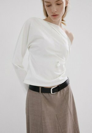 Femme portant un haut blanc asymétrique à manches longues avec une épaule découverte, une jupe plissée beige et une ceinture noire avec boucle argentée.