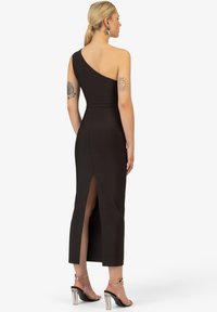 Zwarte een-schouderjurk met een aansluitende silhouet, hoge split tot de knie aan de achterkant en een zachte, rekbare stof. Duidelijke sandalen met blokhak.
