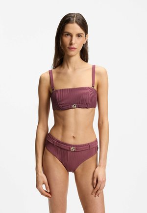 Frau trägt einen malvenfarbenen, gerippten Bikini mit goldenen Schnallendetails am Bandeau-Oberteil und hoch taillierten Höschen, stehend vor weißem Hintergrund.