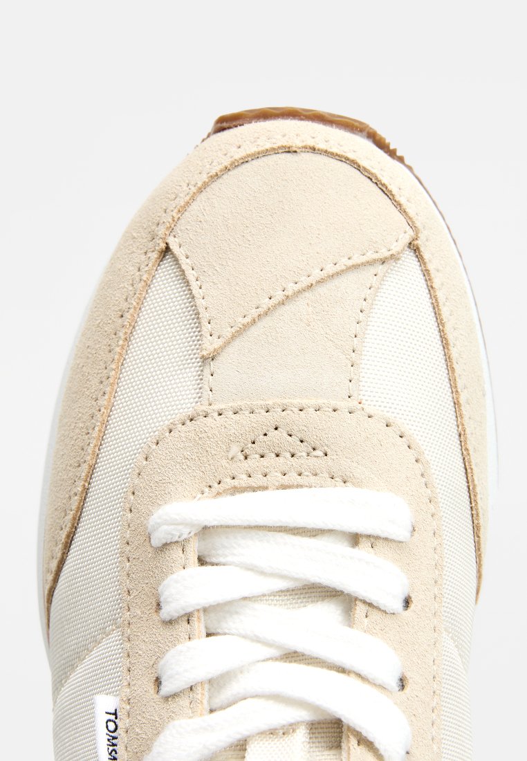 Zapatilla en tonos beige y blanco con puntera de gamuza y paneles de malla, cordones blancos y una pequeña etiqueta de tela en el lateral.