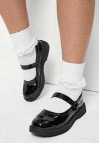 Svarta lackläder Mary Jane-skor med ett säkerhetsrem, tillsammans med vita volangsockor vid ankeln, som visar en texturerad sula.