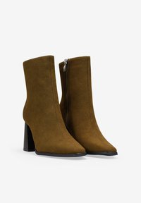 Botas de ante marrón hasta el tobillo con punta afilada y tacón cuadrado grueso. Presentan una cremallera lateral y una textura suave.