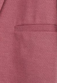 Bordowy materiał marynarki o teksturze z delikatnym diagonalnym wzorem, z jedną kieszenią po lewej stronie i gładkimi detalami szycia.