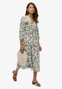 Abito midi a fantasia verde e bianca con scollatura a V, maniche a tre quarti e design a strati, abbinato a una borsa intrecciata beige e tacchi cubani marroni.