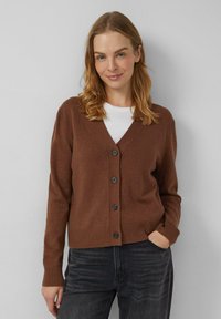 Brauner Cardigan mit V-Ausschnitt, ausgestattet mit fünf schwarzen Knöpfen, gerippten Bündchen und Saum; getragen über einem weißen Oberteil, kombiniert mit dunkelgrauen Jeans.