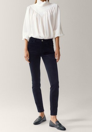 Kvinde iført en hvid bluse med høj hals og puffede ærmer, sorte slim-fit bukser og sorte loafers, stående mod en ensfarvet baggrund.