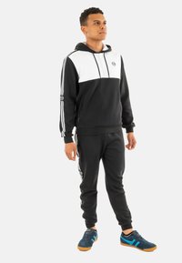 Felpa nera e bianca con zip sul davanti, abbinata a pantaloni jogger neri. Presenta tre strisce bianche sulle maniche e sui pantaloni. Scarpe blu e nere.