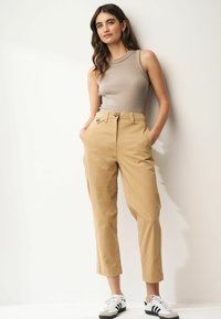 Tan cotton trousers with a high waist, side pockets, and a relaxed fit. Paired with a beige ribbed tank top and black-white sneakers.