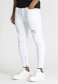 Uske mršave bijele traperice s izrezima, patentnim zatvaračem na gležnju i pet džepova, uparenim s bijelim tenisicama. Mekani denim materijal.