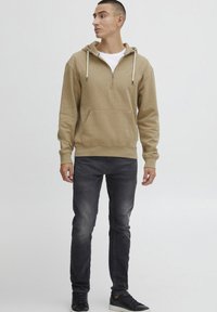Felpa con cappuccio marrone, dotata di tasca frontale, collo con zip e cordini, abbinata a jeans slim-fit scuri e sneakers nere.