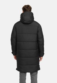 Zwarte pufferjack, lange uitvoering met horizontaal gestikte secties, grote capuchon en elastische manchetten. Gladde textuur, minimale details.