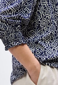 Χέρι σε τσέπη μπεζ παντελονιού, που φοράει μπλε και λευκό πουκάμισο με μοτίβο και μακριά μανίκια με ελαστική μανσέτα.