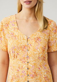 Vestido floral amarillo con mangas cortas abullonadas, escote en V y frontal de botones con pequeños botones grises. Patrón floral en rosa y blanco.