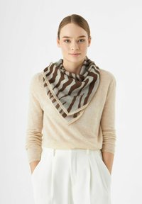 Bruine en lichtgrijze sjaal met een gestreept patroon, in een driehoekige vorm gelaagd, gedragen over een beige trui en witte broek.