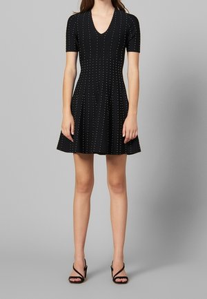 Femme portant une robe courte noire à col en V avec de petits pois blancs, assortie à des sandales noires à talons hauts.