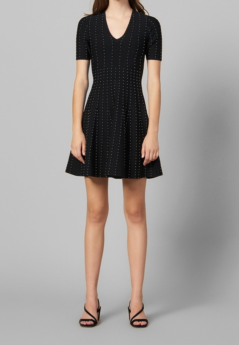 Femme portant une robe courte noire à col en V avec de petits pois blancs, assortie à des sandales noires à talons hauts.