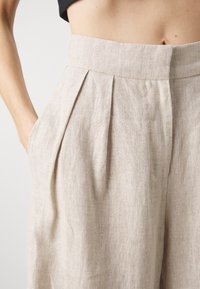 Ljusbeige linbyxor med veck, hög midja, sidofickor och en texturerad yta som visar upp ett naturligt, andningsbart material.