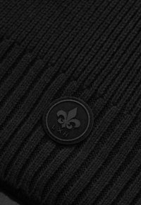 Schwarze gestrickte Mütze mit gerippter Textur, die ein kreisförmiges, geprägtes Logo mit floralem Design und "LXH" am unteren Rand zeigt.