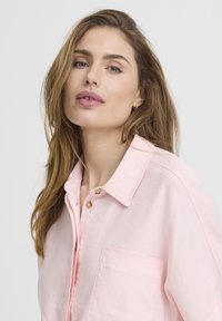 Femme aux longs cheveux châtain clair portant une chemise ample rose pâle à boutons, regardant légèrement vers le haut sur un fond blanc uni.