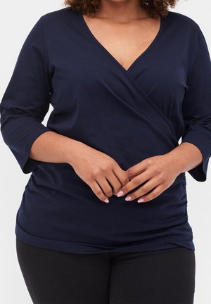 Femme portant un haut cache-cœur bleu marine à col en V avec manches trois-quarts et un pantalon noir, les mains jointes devant, de face.