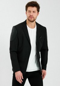Zwarte blazer met geribbelde leren schouderaccenten, sluiting met één knop en nepzakdetail, gedragen over een witte T-shirt en zwarte broek.