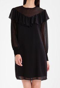 Femme portant une robe noire avec des manches longues transparentes et un détail volanté sur le plastron au niveau de la poitrine et des épaules.