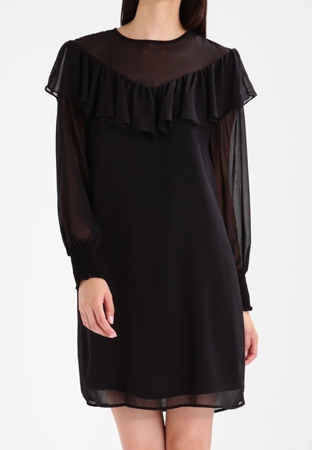 Femme portant une robe noire avec des manches longues transparentes et un détail volanté sur le plastron au niveau de la poitrine et des épaules.