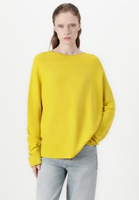 Kollane, oversized kudum spetsiifne, millel on laiem kaelus ja pikad varrukad. Kanga tekstuur on pehme. Kombineeritud helesiniste teksadega.