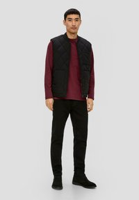 Czarna pikowana kamizelka na maroonowej koszuli z długim rękawem, w połączeniu z czarnymi spodniami i czarnymi botkami za kostkę. Prosty, funkcjonalny design.