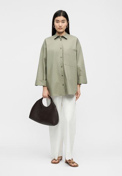 Γυναίκα όρθια, φοράει φαρδιά ανοιχτοπράσινη πουκαμίσα με κουμπιά, λευκό παντελόνι φαρδύ με κοψίματα στο τελείωμα, ταμπά σανδάλια και κρατάει μια μεγάλη σκούρα καφέ τσάντα.