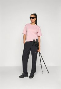 Gaiši rozā īss piedurkņu krekls un melnas kravas bikses, aprīkotas ar pārgājienu nūjām un tumšām kurpēm. Vienkāršs dizains ar praktiskiem rāvējslēdzējiem.