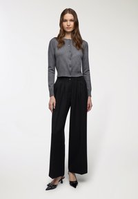 Giovane donna che indossa un cardigan grigio abbottonato, pantaloni neri a gamba larga e décolleté nere con fiocchi su uno sfondo chiaro e semplice.