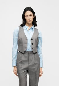 Γκρι γιλέκο με τρία μαύρα κουμπιά, φορεμένο πάνω από ένα ανοιχτό μπλε πουκάμισο με κουμπιά. Το γιλέκο έχει εφαρμοστή γραμμή με ελαφριά γυαλάδα.