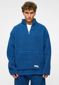 Blauwe fleece trui met ritssluiting, voorzak en textuur. Losvallend ontwerp met een label op de onderste zoom.