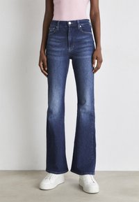 Jeans acampanados de color azul oscuro, hechos de denim, con una cintura alta, un ligero desgaste y un clásico diseño de cinco bolsillos, combinados con zapatillas blancas.