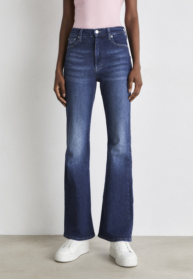 Jeans acampanados de color azul oscuro, hechos de denim, con una cintura alta, un ligero desgaste y un clásico diseño de cinco bolsillos, combinados con zapatillas blancas.