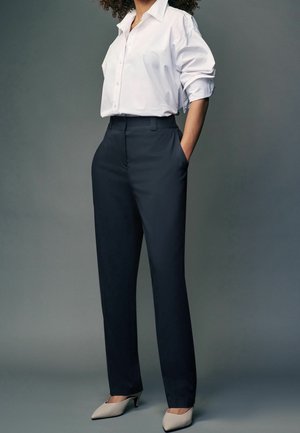 Woman wearing a white button-up shirt tucked into high-waisted black trousers, with hands in pockets and beige pointed heels.