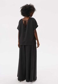 Zwarte jumpsuit met wijde pijpen en korte mouwen, met een diepe V-hals aan de achterkant met striksluiting. Soepele, vloeiende stof met een relaxte pasvorm.