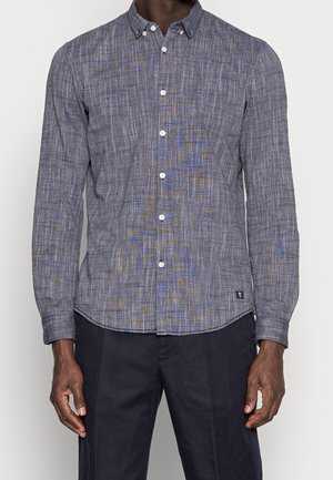 Mężczyzna ubrany w długorękawową koszulę na guziki z niebiesko-szarym teksturowanym wzorem oraz ciemne spodnie, stojący na jednolitym tle.