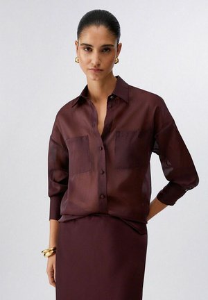 Donna con capelli tirati indietro, che indossa orecchini a cerchio d'oro, braccialetti d'oro, una camicia trasparente color bordeaux con bottoni e una gonna abbinata su sfondo bianco.