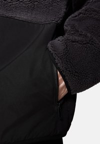 Czarna i szara polarowa kurtka z zamkiem błyskawicznym, kieszenią na zamek, chropowatym górnym materiałem i gładką dolną częścią z kontrastujących materiałów.